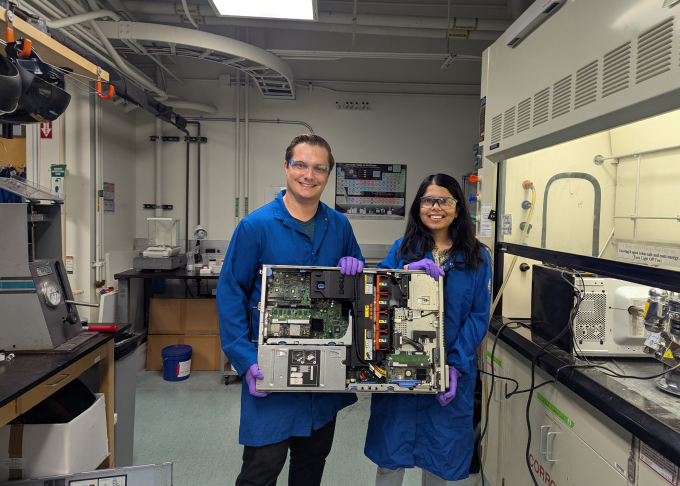 Two UCSB researchers wearing blue coats stand in a laboratory holding a circuit board